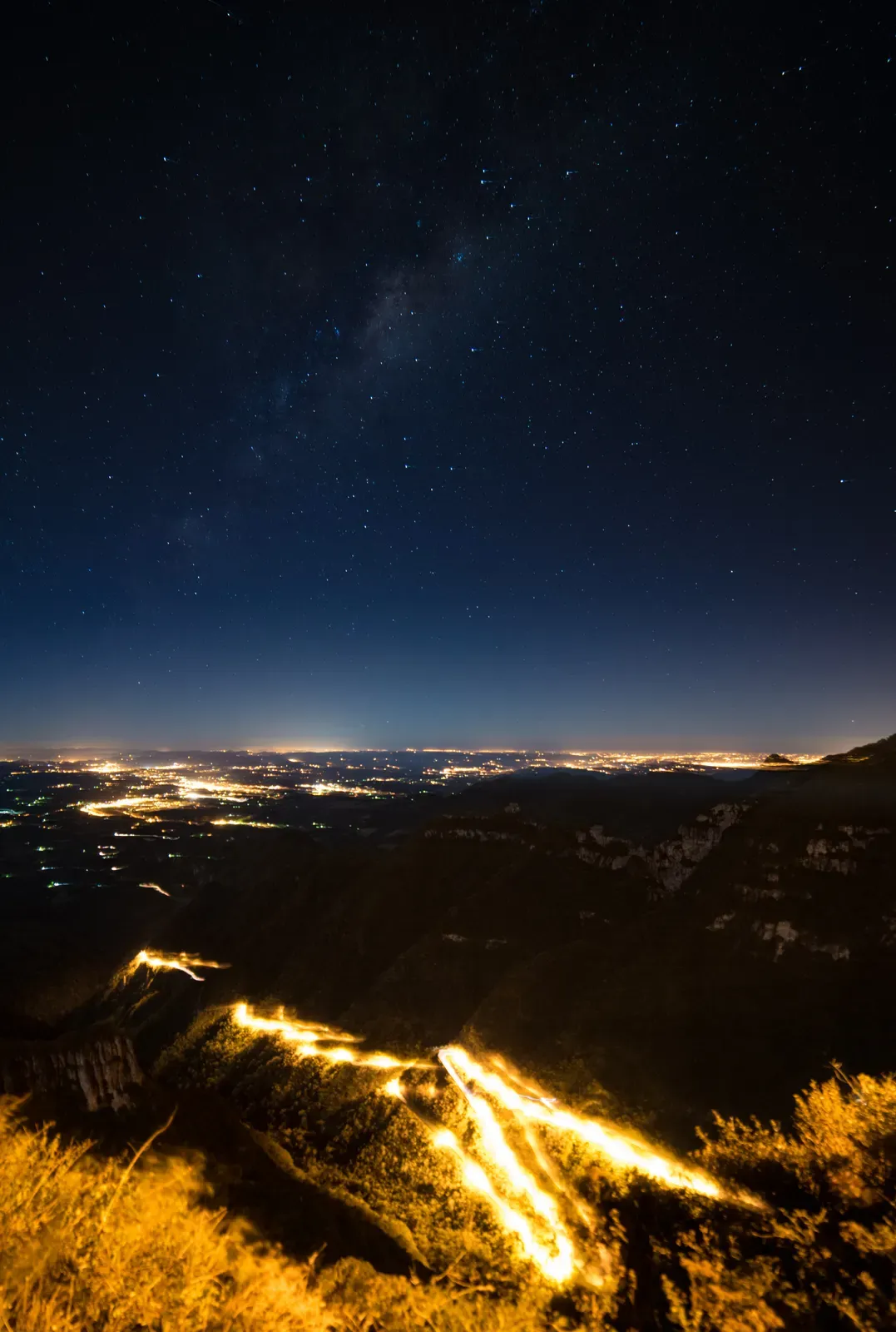 Vista noturna do alto da serra, com as luzes das cidades do litoral espalhadas na planície e o céu estrelado